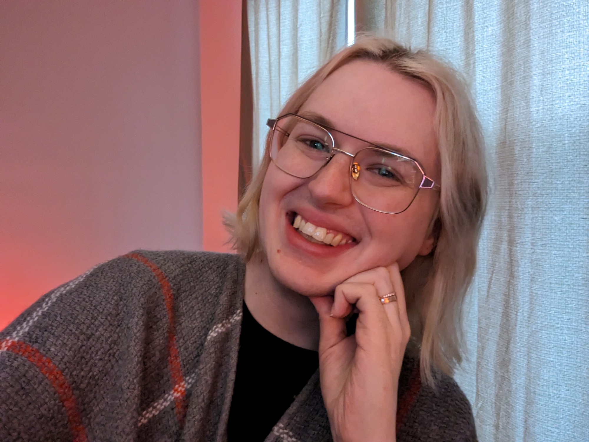 Blonde woman sitting at her desk wearing a gray poncho with red and white stripes, smiling into camera. She is leaning on her hand, wedding rings are visible.