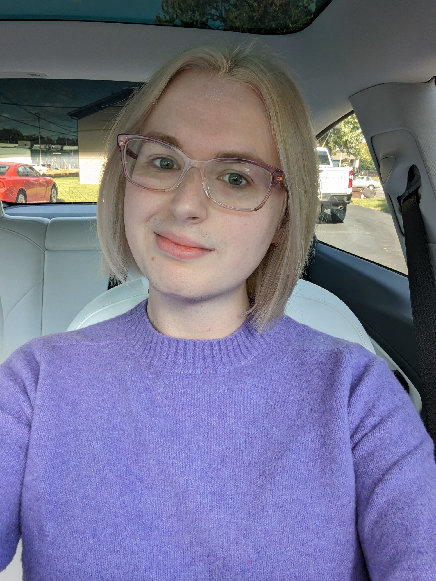 Blonde woman sitting in car, wearing purple sweater. Hair is chin-length and straight, newly bleached and dyed.