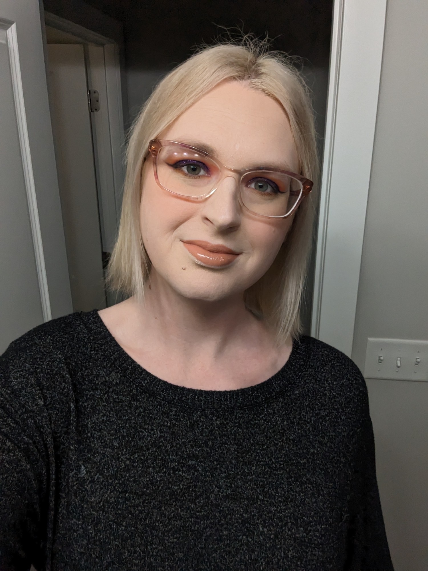 sky looking at the camera with a slight smile, she has on makeup with sunset-themed eyeshadow, orange blush, and a peach lipstick with gloss. she has slightly shorter than shoulder-length straight blo