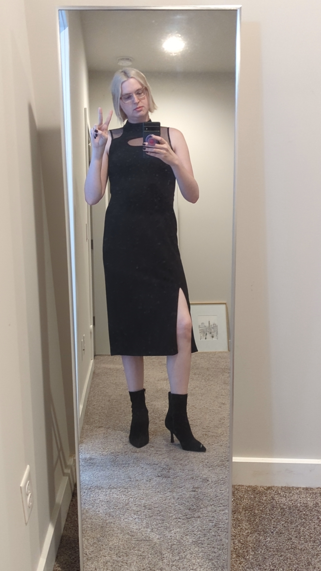 Sky standing in front of a full-length mirror, making a peace sign. She's wearing a black dress that goes just past her knees, and has mesh above the breast. She is wearing black suede heel boots.