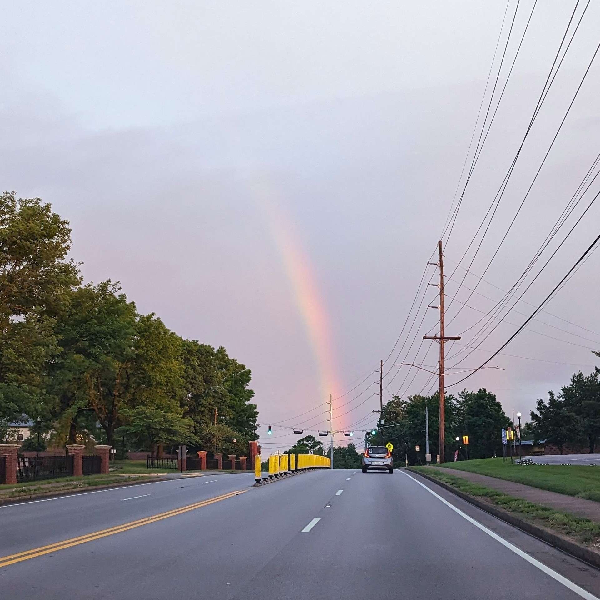 the end of a rainbow, going into a road with yellow bollards in the middle. a car is driving away from the camera and there's a sidewalk and power lines on the right
