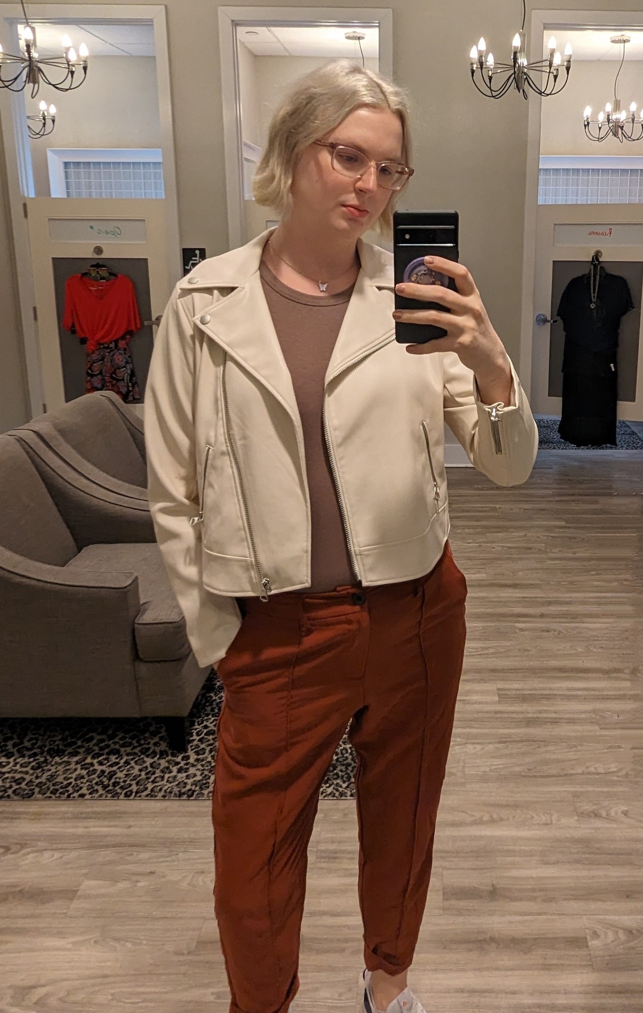 tall blonde woman taking a selfie in a mirror at a Torrid dressing room. She's wearing burnt orange pants, a brown bodysuit, and a light tan leather jacket.
