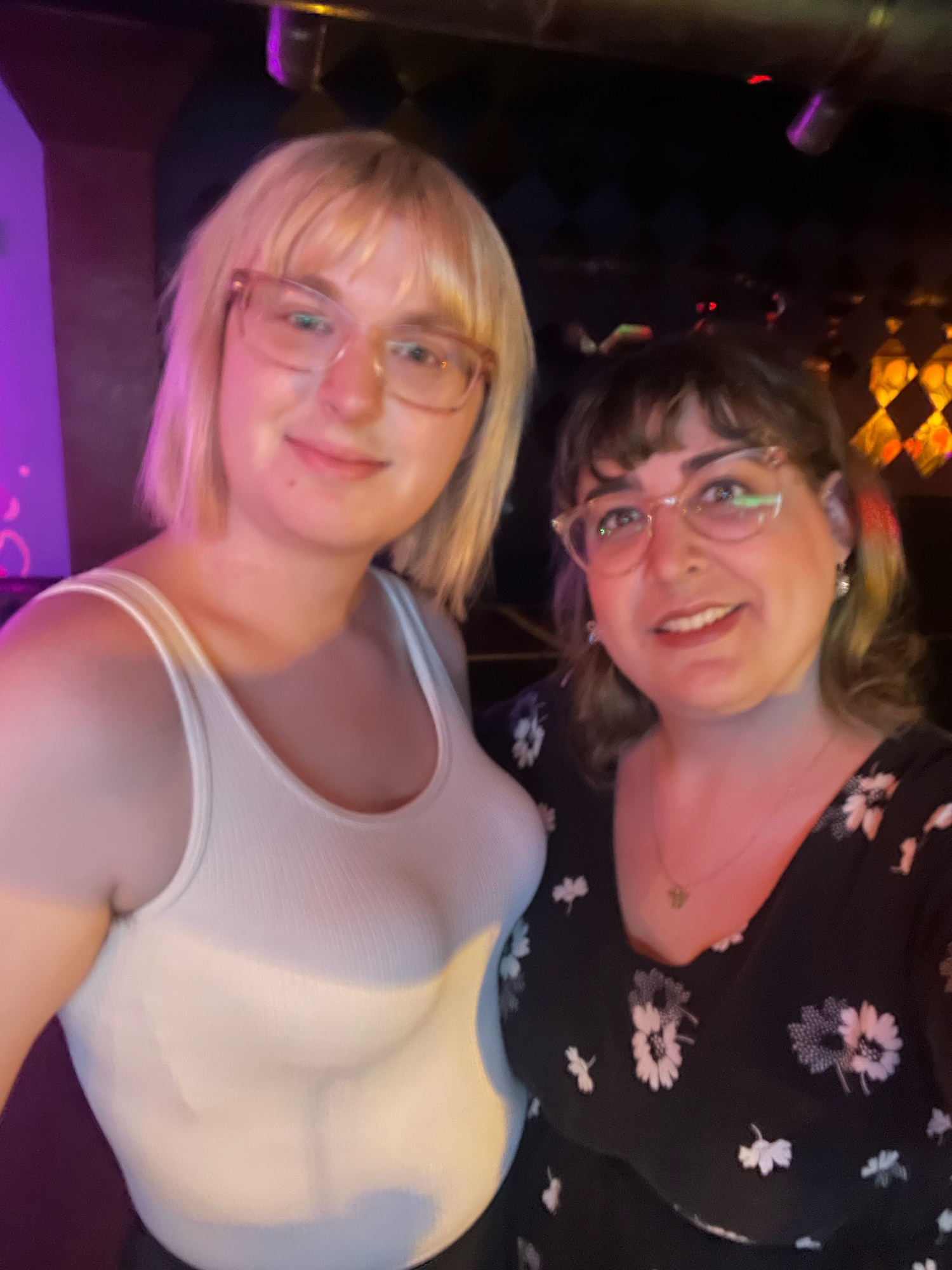 a selfie of skylar and her wife olivia at a bar. skylar is a blonde woman with short hair and bangs, wearing glasses and a white tank top. olivia is a brunette woman with green highlights and nose pie