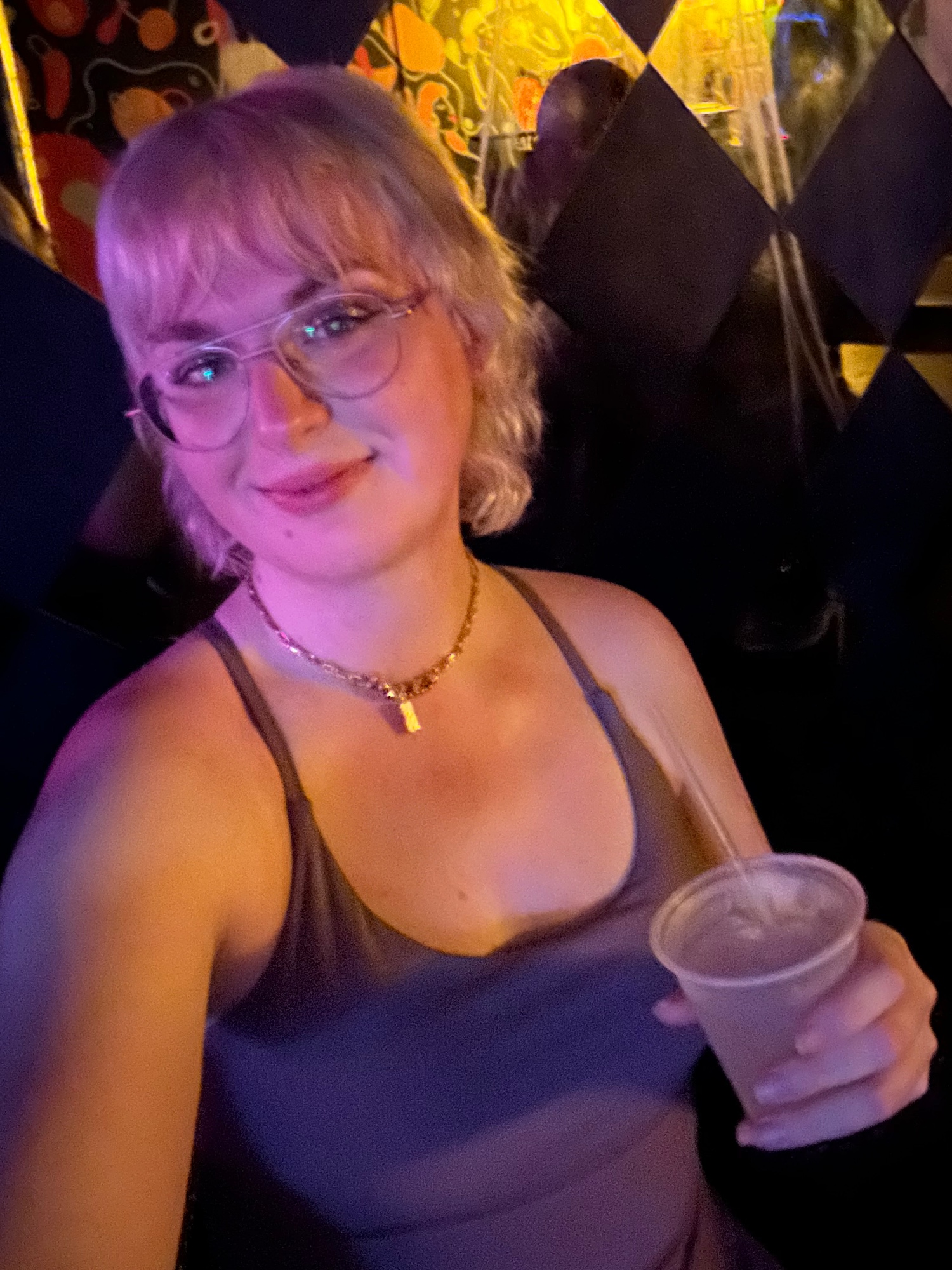 a blonde woman with bangs sits in bar lighting, she’s wearing a dark purple dress and holding a cocktail.