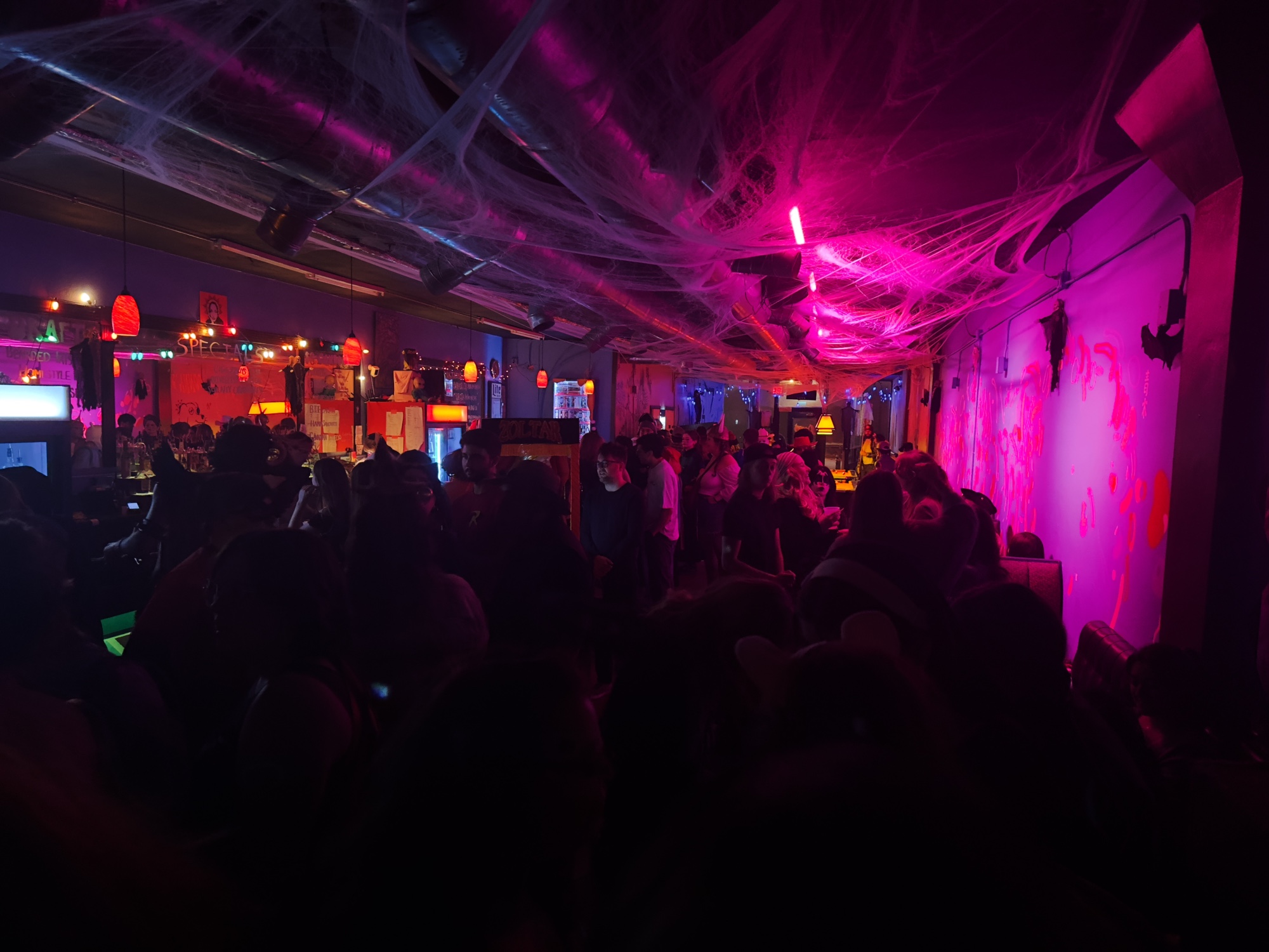 a dark photo of a bar packed with patrons; there’s hot pink lighting on the right side, and the ceiling is covered in fake spider webs for halloween.