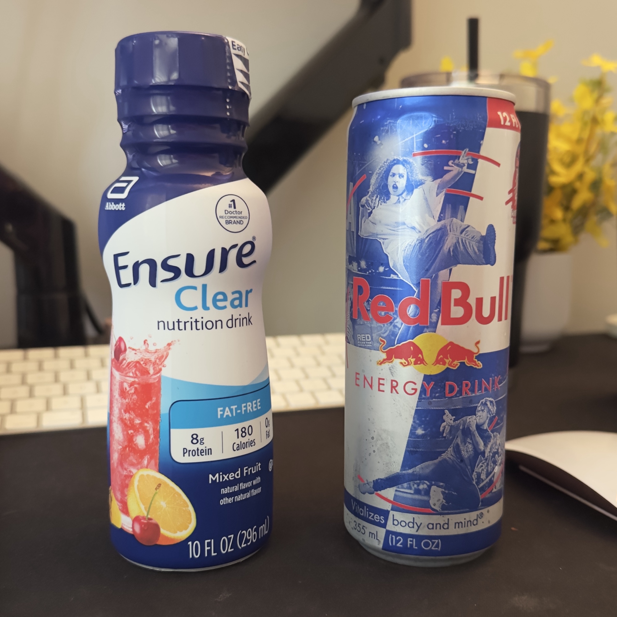 a bottle of ensure clear mixed fruit flavor sitting to the left of a can of original red bull on a desk, on a black mousepad. there’s a keyboard behind them and a mouse to the right. an artificial yel