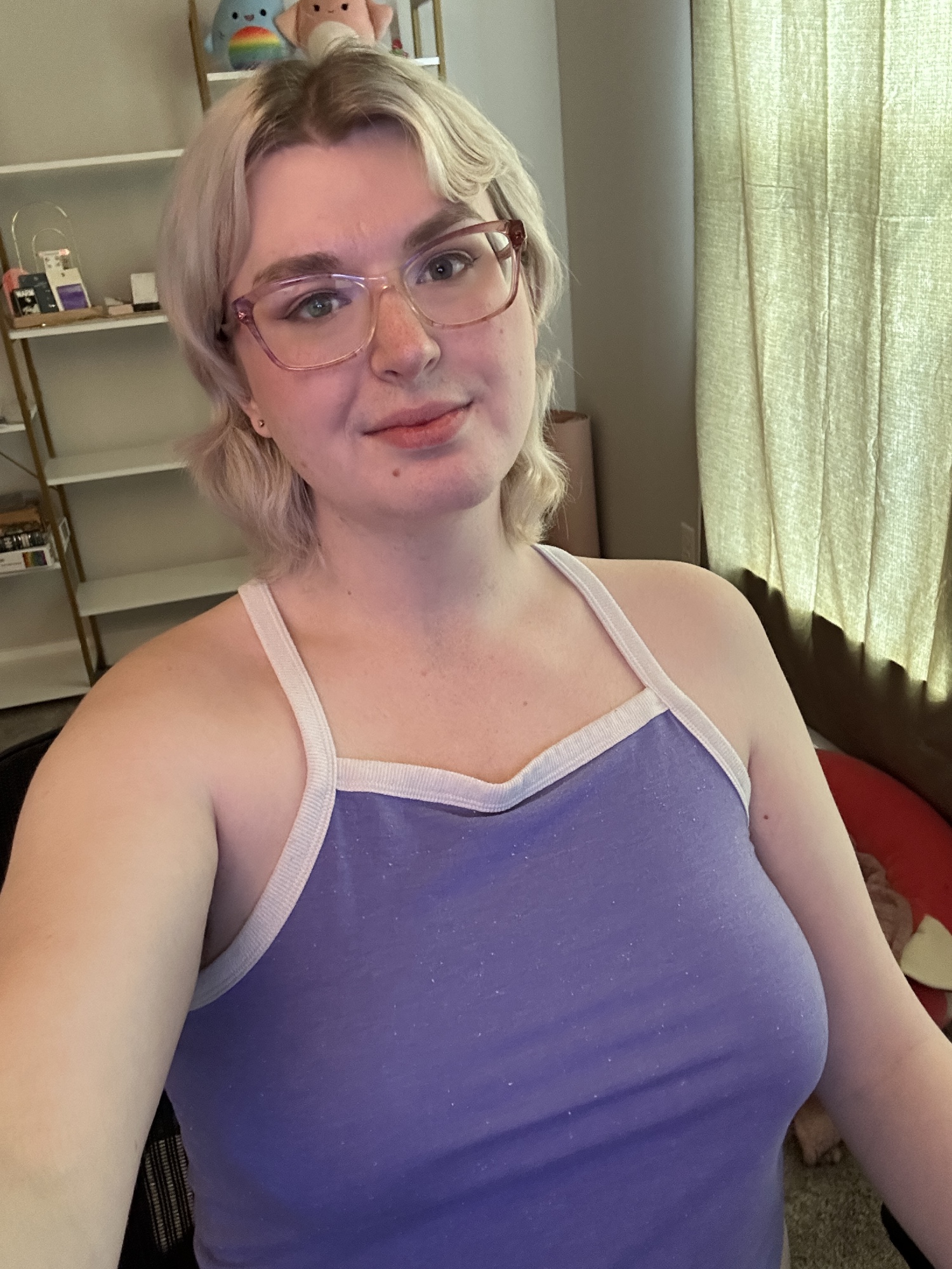 a photo of zari, sitting at their desk, wearing a purple tank top. they have short-ish blonde hair with noticeable roots, and large pink plastic glasses.