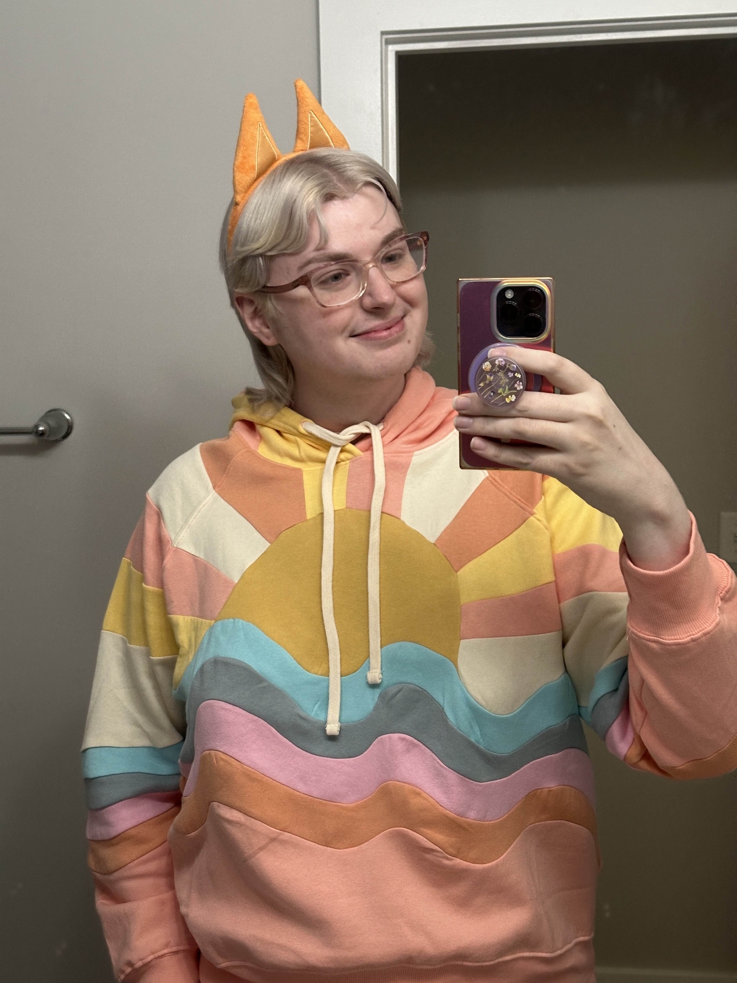 a mirror selfie of zari, wearing a hoodie that has pink and orange wavy lines of fabric at the bottom, then a blue wave, a sun coming out of that, and white, pink, and yellow stripes coming out of the