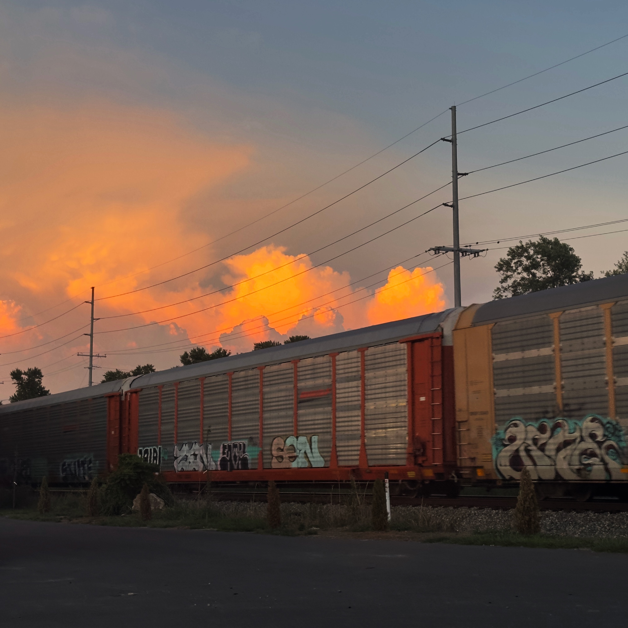 a train, in the center of the frame is a car with orange and silver metal, with graffiti along the bottom. in the background is a deep orange sunset shining through clouds, and there are power lines i