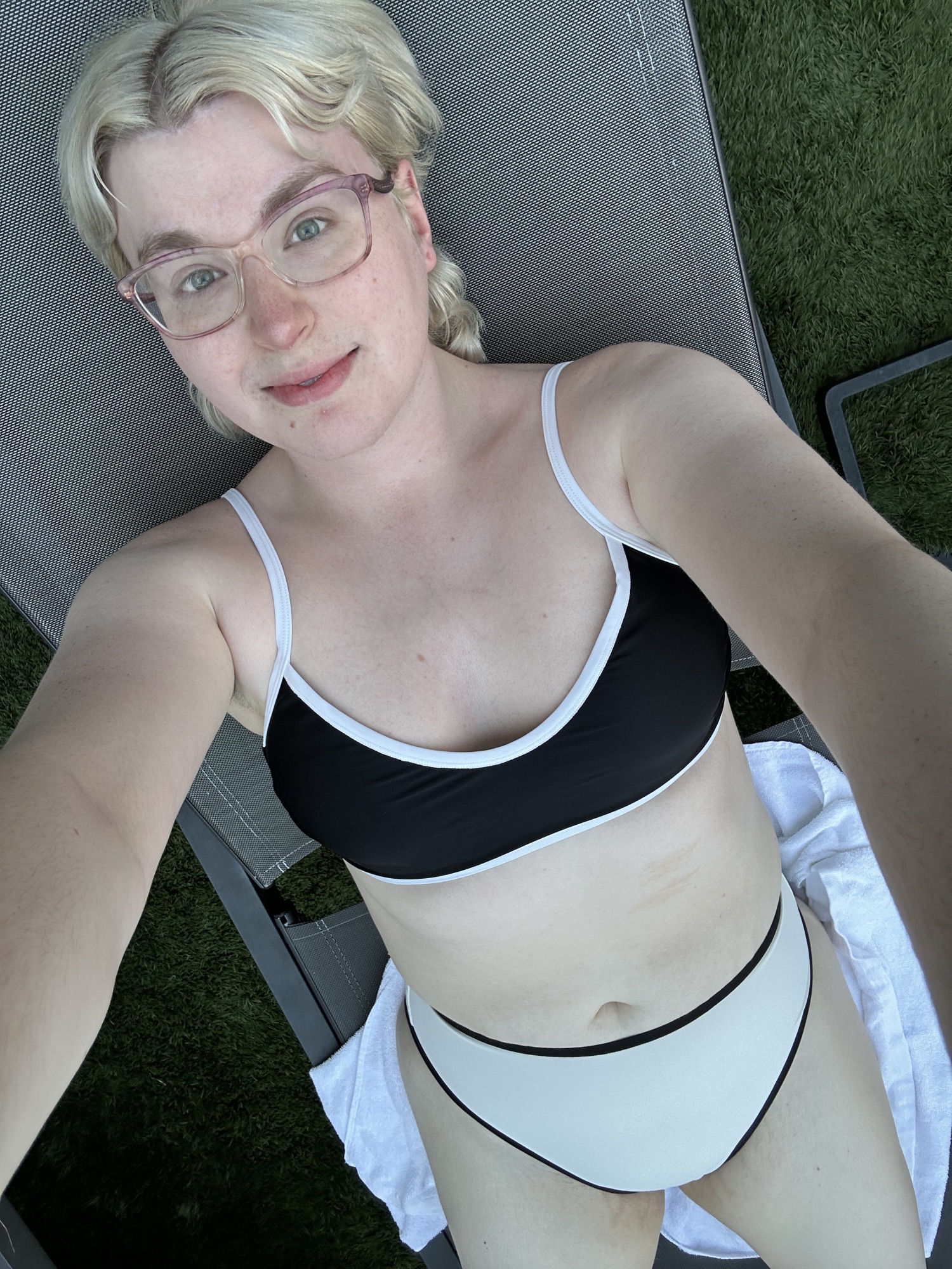 zari laying on a lounge chair by the pool, the chair is sitting on top of fake grass. zari is wearing a bikini, the top is navy with cream piping, the bottoms are cream with navy piping. they have sho