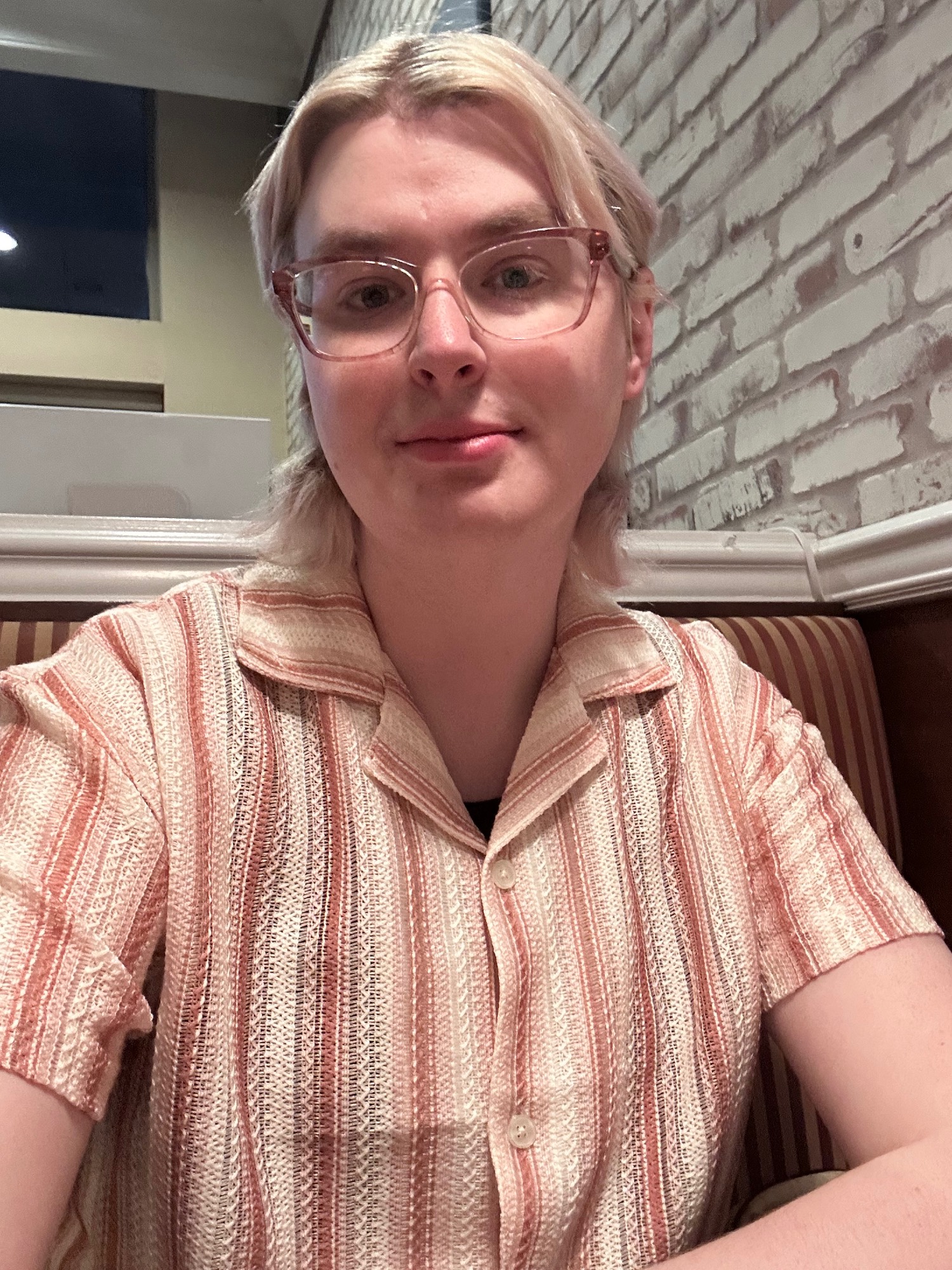 a selfie of ethan sitting at a booth in a restaurant, wearing a linen button-up short sleeve shirt with a collar with dark pink, light tan, and white vertical stripes in a knit type pattern. he has sh