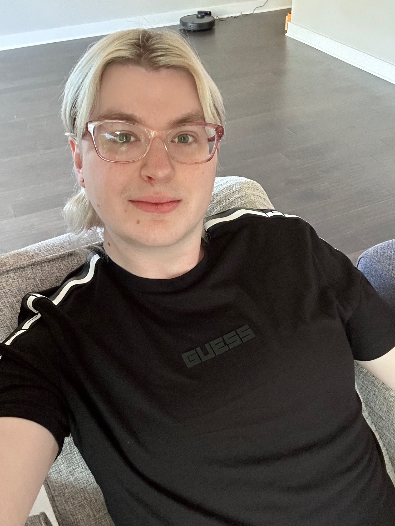 a selfie of ethan sitting in a recliner, wearing a black t-shirt with white stripes on the shoulders and a black rubber "GUESS" logo. he has short, shoulder length blonde hair with visible roots that'