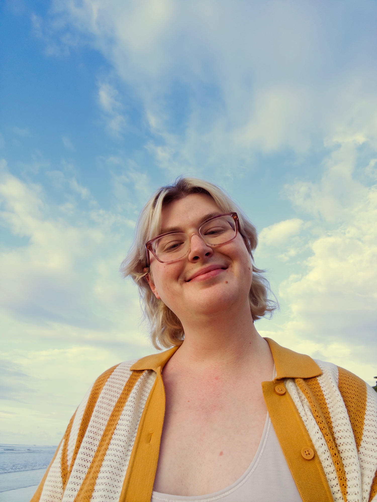 a low-angle selfie of skylar smiling, with short blonde hair and glasses, wearing a mustard yellow and white striped cardigan against a bright blue sky with clouds