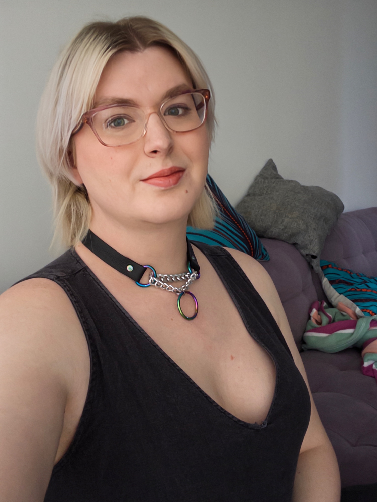 skylar, a woman with blonde hair and glasses wearing a black romper and a black leather martingale collar with rainbow ring. they are sitting on a purple couch with a teal pillow in the background.