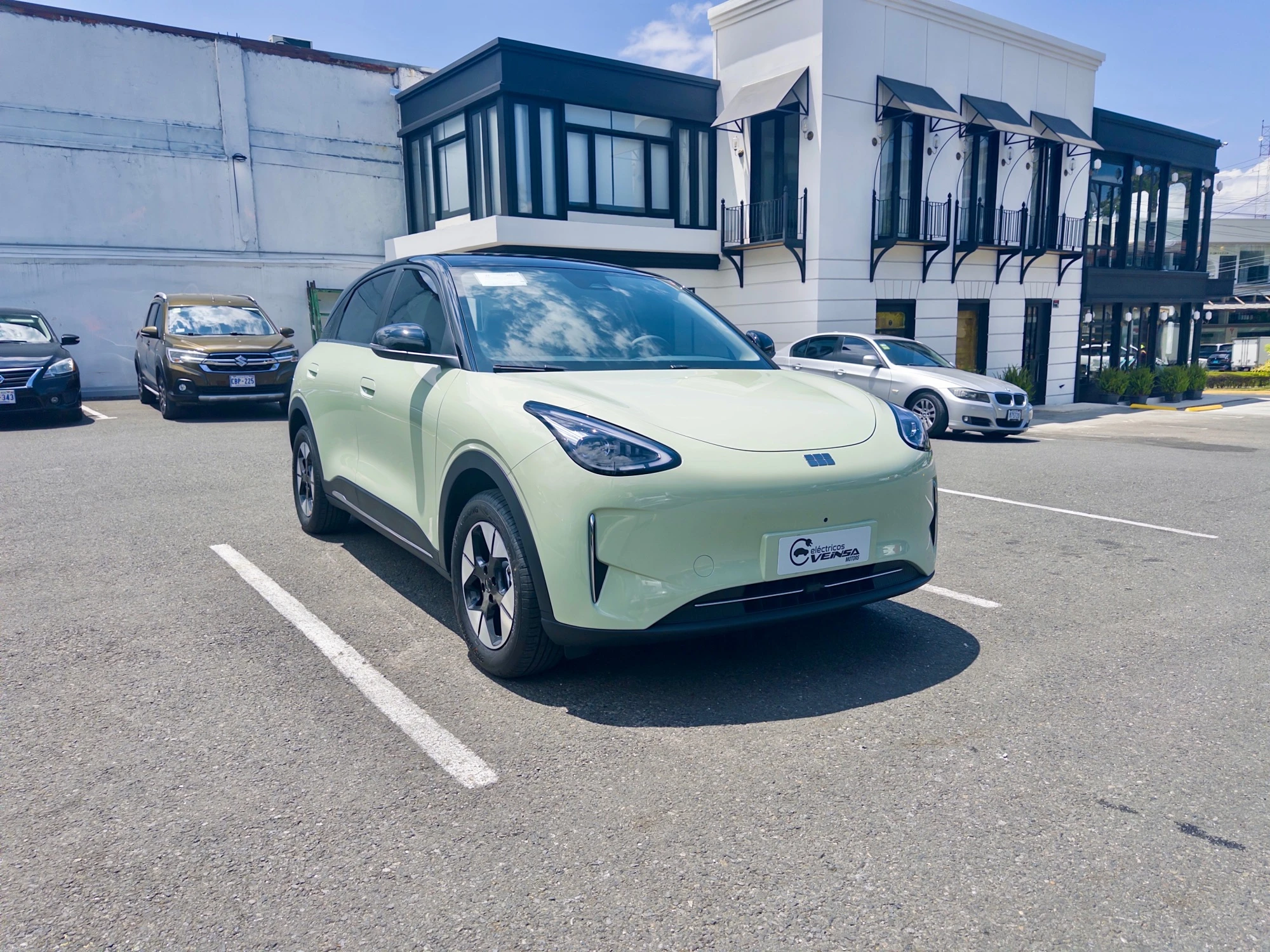 A light green Geely Geome GT+ (EX2) electric crossover parked in a parking lot in San José, Costa Rica. The compact SUV features a smooth, rounded front fascia with split headlights, two-tone black roof, and distinctive angular alloy wheels. A Veinsa dealer plate is visible on the front. Other vehicles and a white building with black-framed windows are in the background under a clear blue sky.
