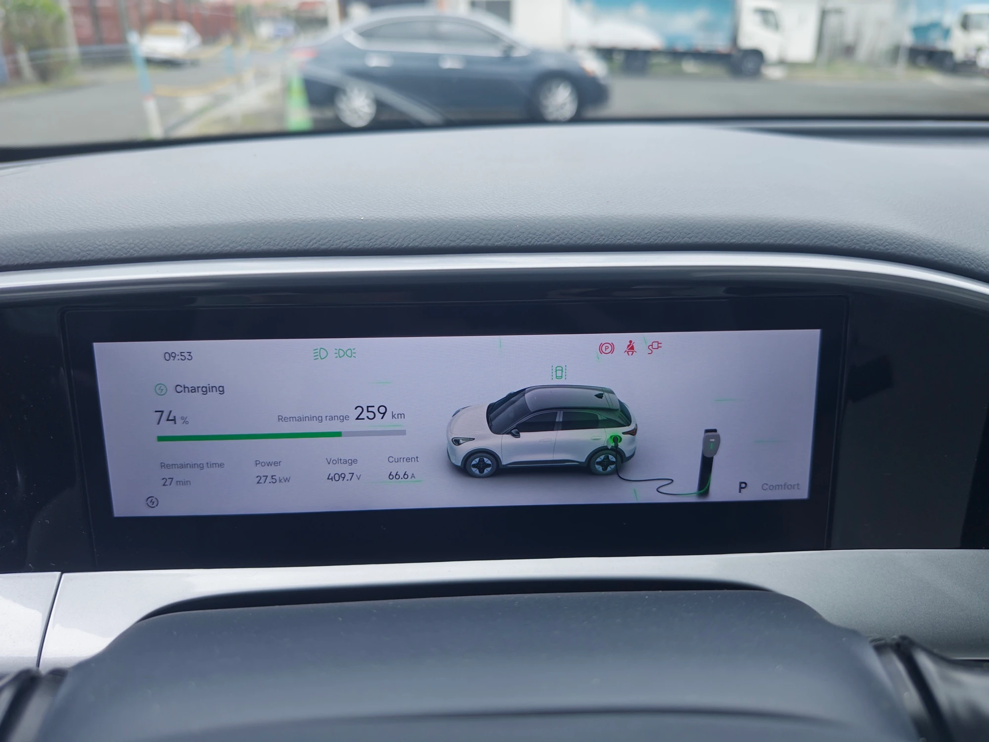 The interior of a Geely EX2 dashboard showing the display while charging. The screen indicates a battery level of 74%, a remaining range of 259 km, and a charging power of 27.5 kW. A digital graphic shows a white and black compact SUV plugged into a charging station. The time is 09:53, and the estimated remaining time to a full charge is 27 minutes.